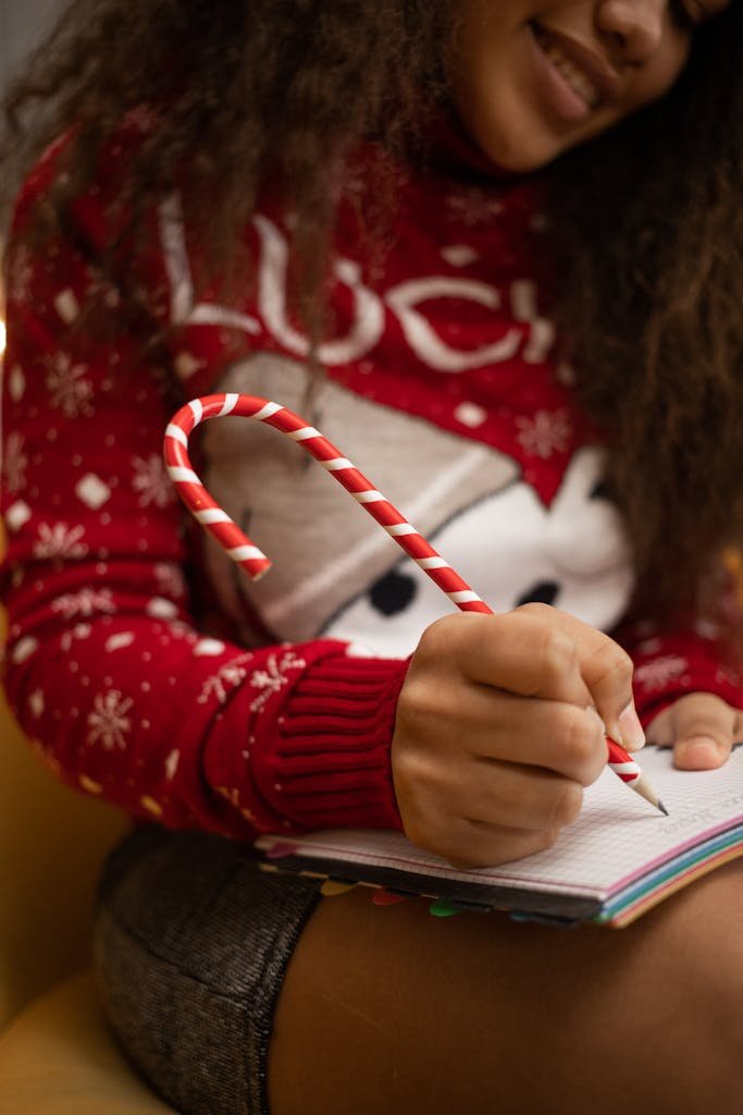 Close-up of a woman writing in a notebook, holding a candy cane pen, wearing a Christmas sweater.